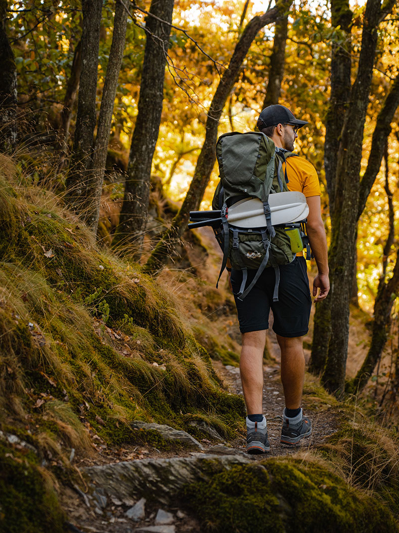 man hikes along hillside forest path with packraft and paddle in his pack