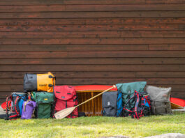 a selection of canoe packs and barrel harnesses in front of a red wood-canvas canoe and wood slat wall