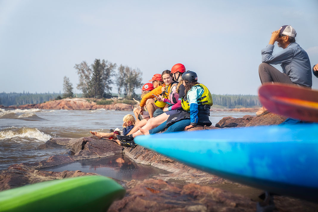 a group of kids and a man sit near kayaks on the banks of the Slave River