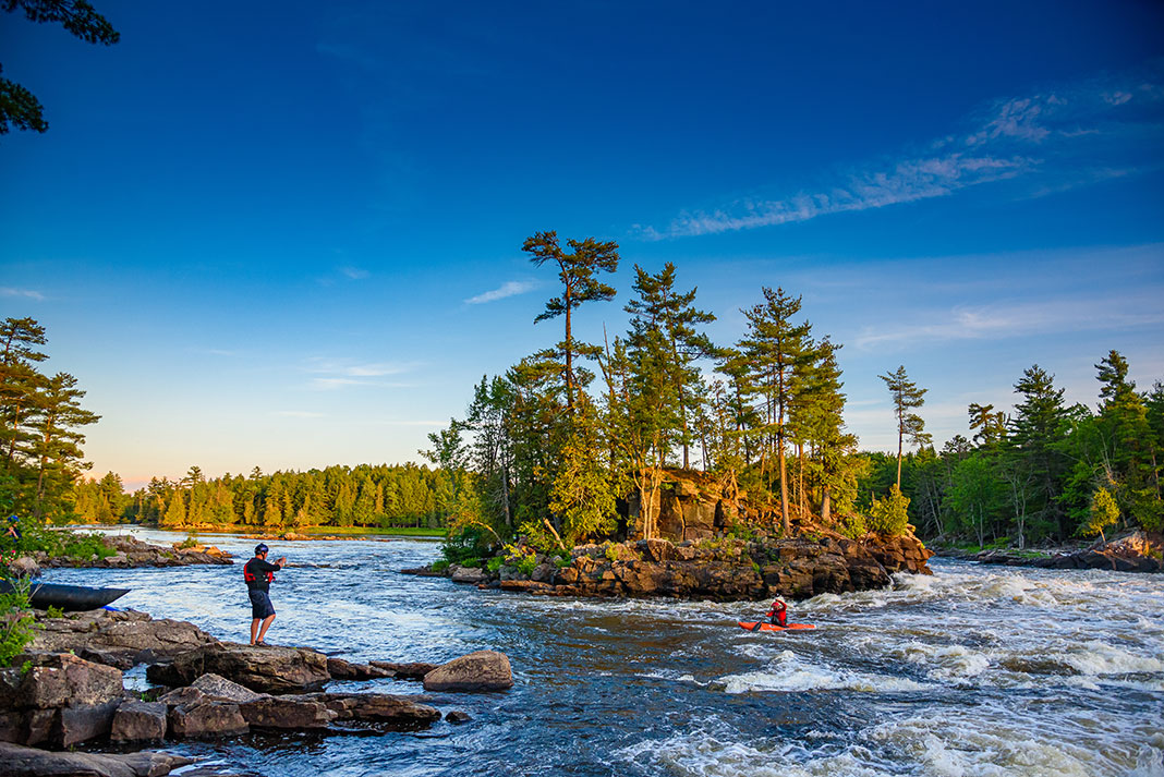 a person on the bank watches while a whitewater kayaker paddles in rapids in front of a small river island