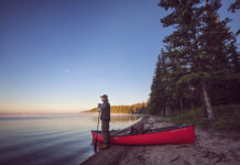man stands at lake's edge holding a paddle beside a tripping canoe at dawn