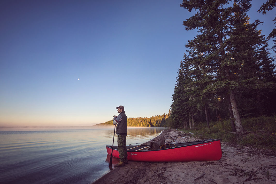 man stands at lake's edge holding a paddle beside a tripping canoe at dawn