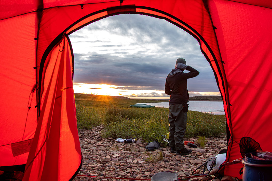 looking out the entrance of a red tent at a camper standing by the river at dawn