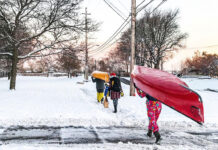 three people carry Christmas gifts and canoes along a snowy sidewalk