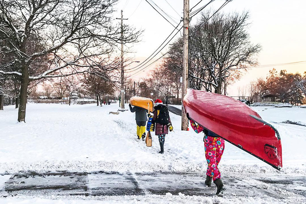 three people carry Christmas gifts and canoes along a snowy sidewalk
