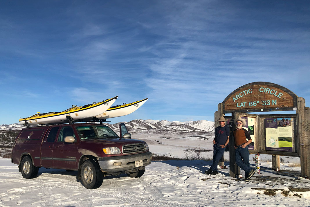 two men stand beside the Arctic Circle near a truck with two sea kayaks on the roof