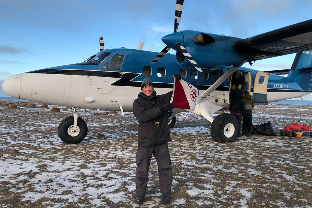 man holds up flag while posing in front of small plane in the Arctic