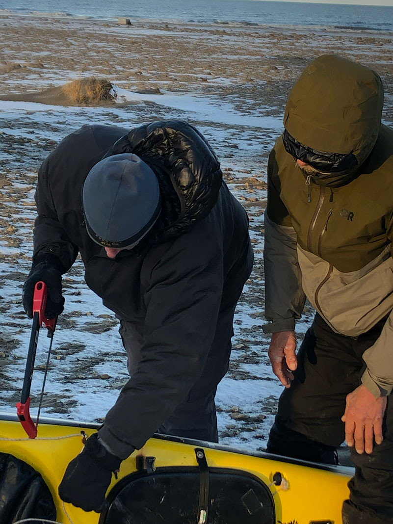 man cuts sea kayak with a hacksaw while standing on the Arctic tundra