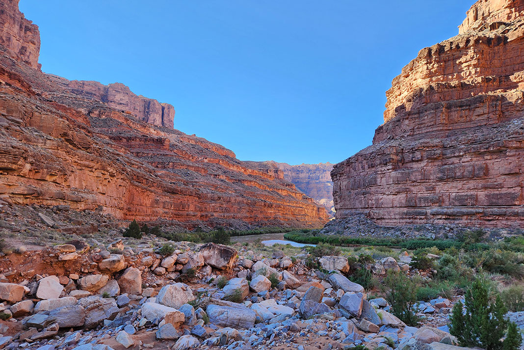 rocks and sparse plants line the banks of the San Juan River, surrounded by steep striated cliffs