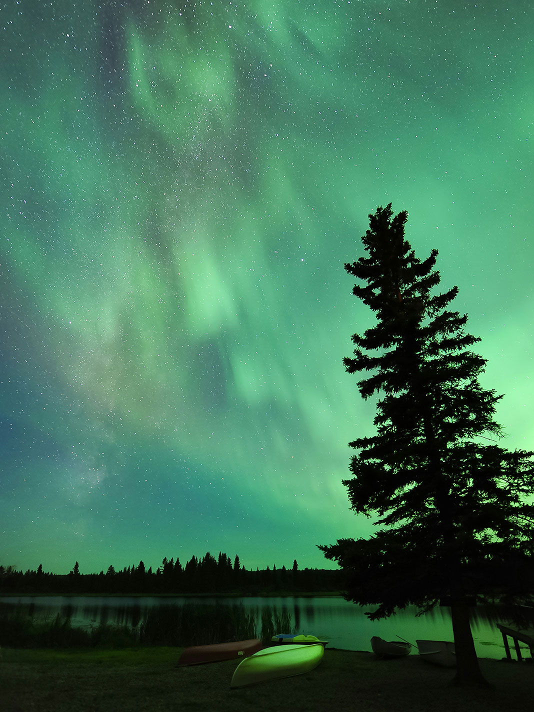 an overturned canoe sits on the bank at a campsite under a vivid night sky with northern lights
