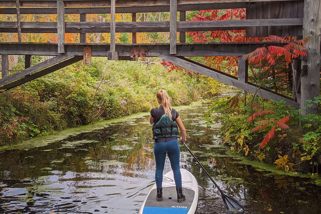 a woman paddleboards underneath a small wooden bridge in the Outaouais region of Quebec