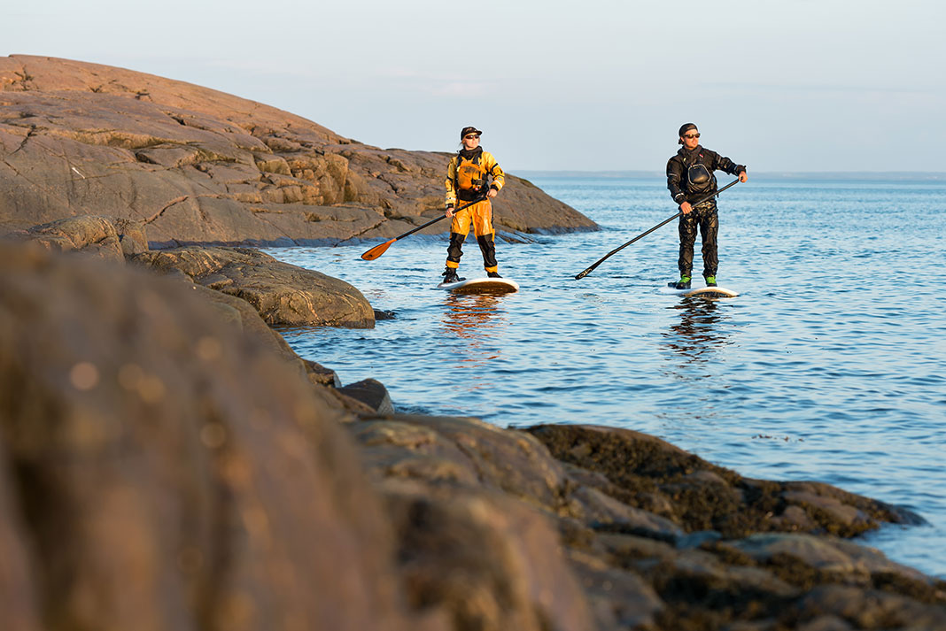 man and woman in drysuits paddleboard along a line of rock caps