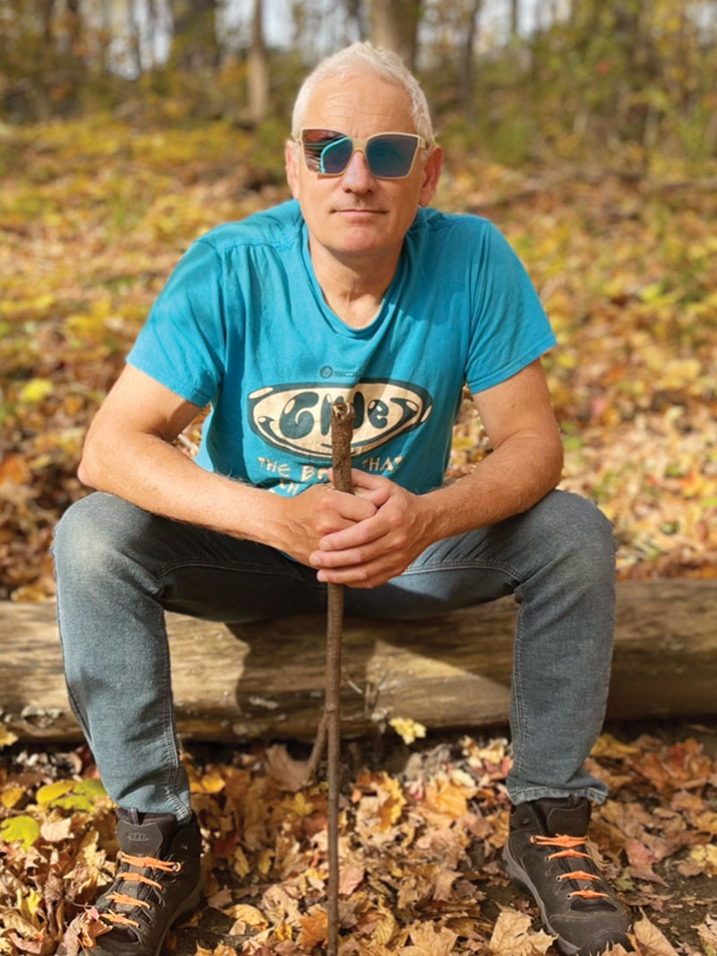 Corran Addison sits on a log while wearing sunglasses and holding something