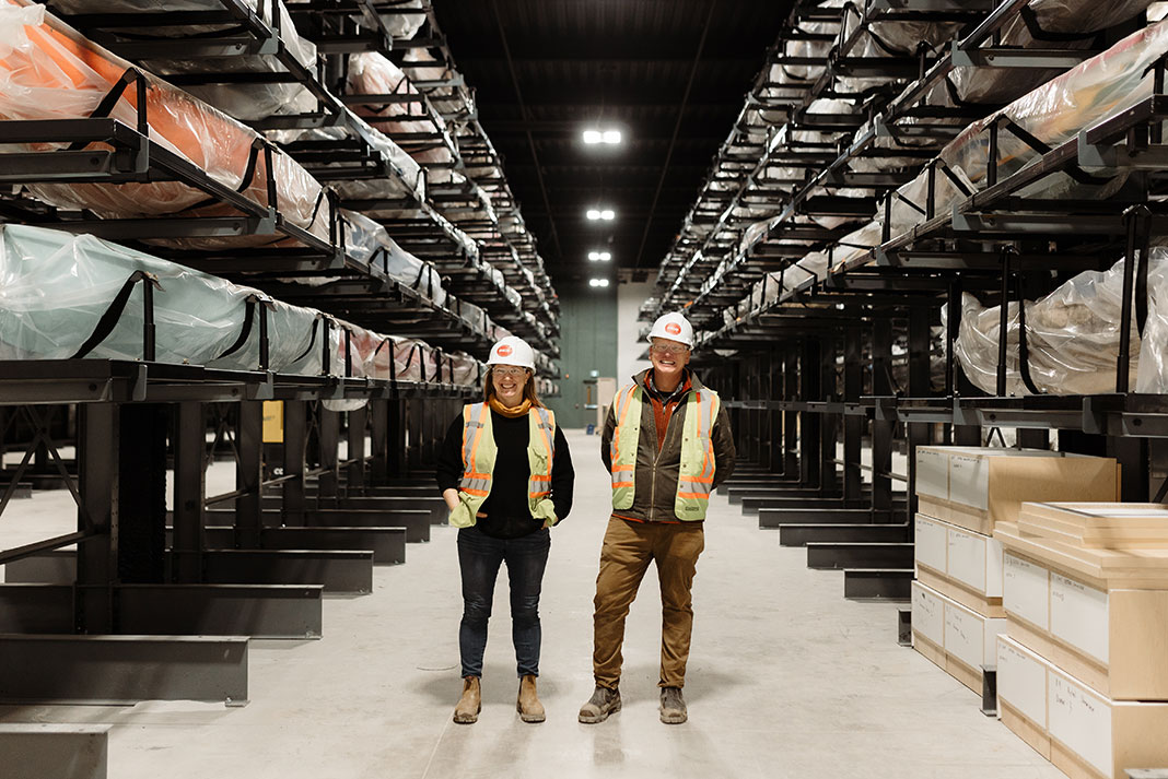man and woman wearing hi-vis vests and white hardhats stand in a storage room at the new Canadian Canoe Museum