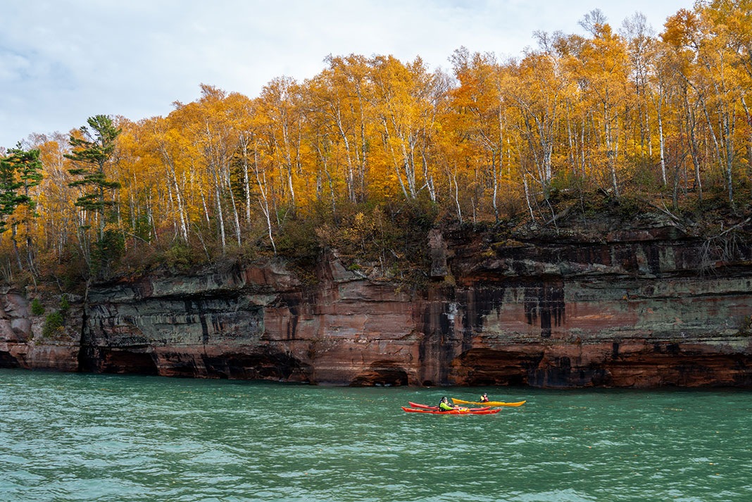 people in a tandem kayak paddle past a rock wall with birch trees on top in Wisconsin