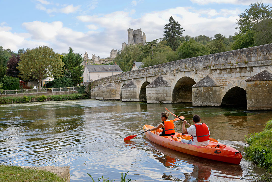 two people paddle a tandem kayak near an old bridge in France