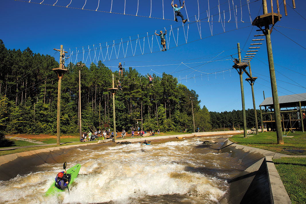a person kayaks through a course at a whitewater park while people climb through a ziplining course above
