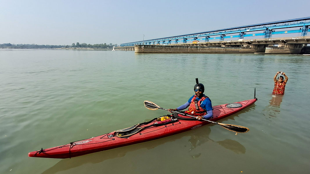 Man poses in touring kayak in India's Ganges River while woman anoints herself with water in the background