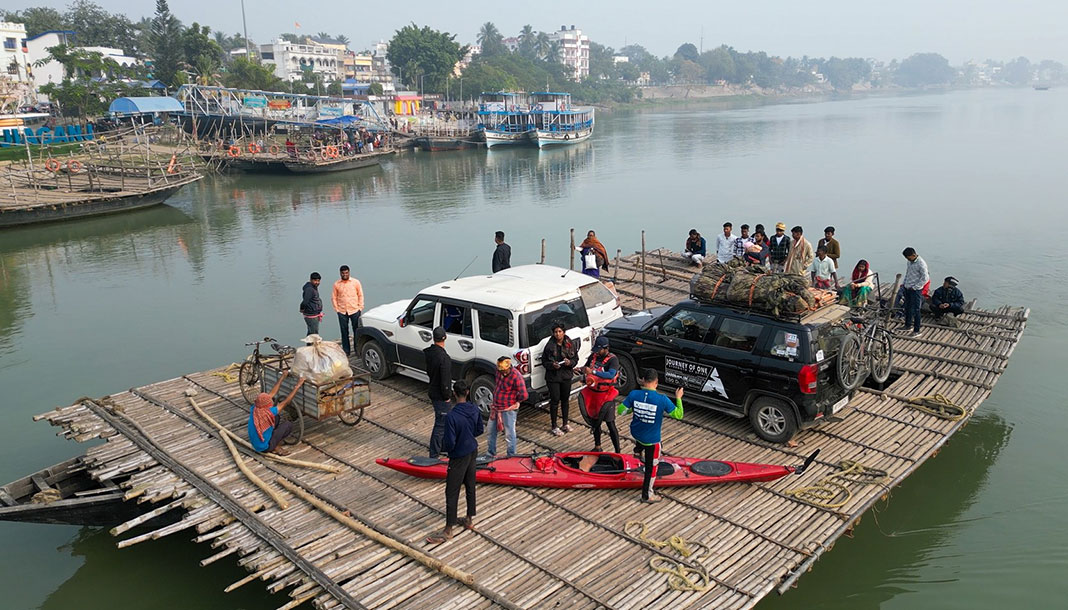 Several vehicles and a kayak join a group of people on a barge floating on the Ganges River