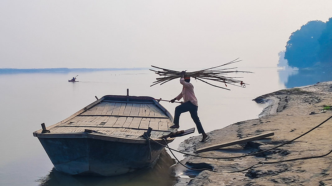 man loads sticks into boat at dawn while kayak paddles in background in India