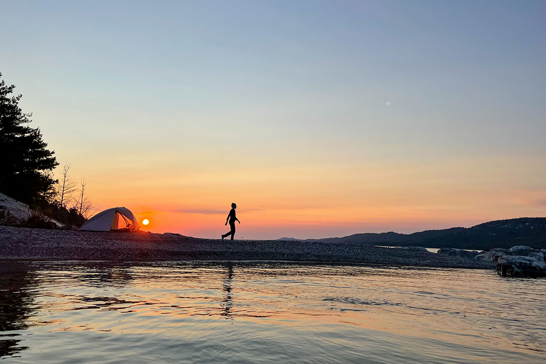 a woman walks around her waterside campsite at dusk while on a microadventure