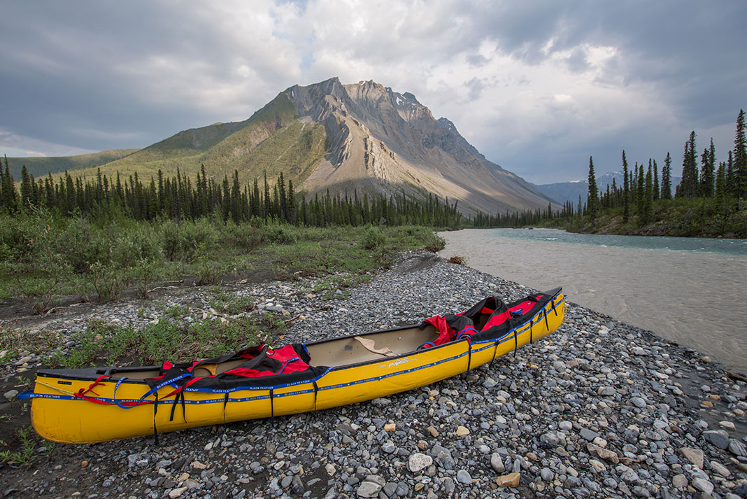 an expedition canoe sits on a pebbly Northwestern Territories riverbank in sumer with dramatic mountain in background