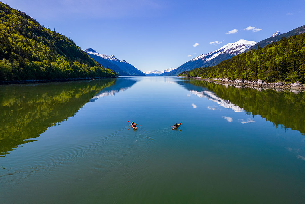 two kayakers sit on placid waters surrounded by mountains while on a summer paddling trip in Alaska