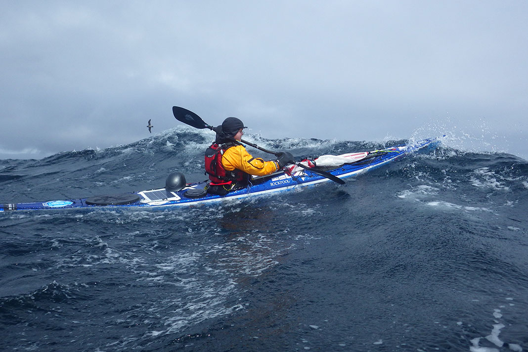 a woman paddles her expedition kayak through ocean swells under grey skies