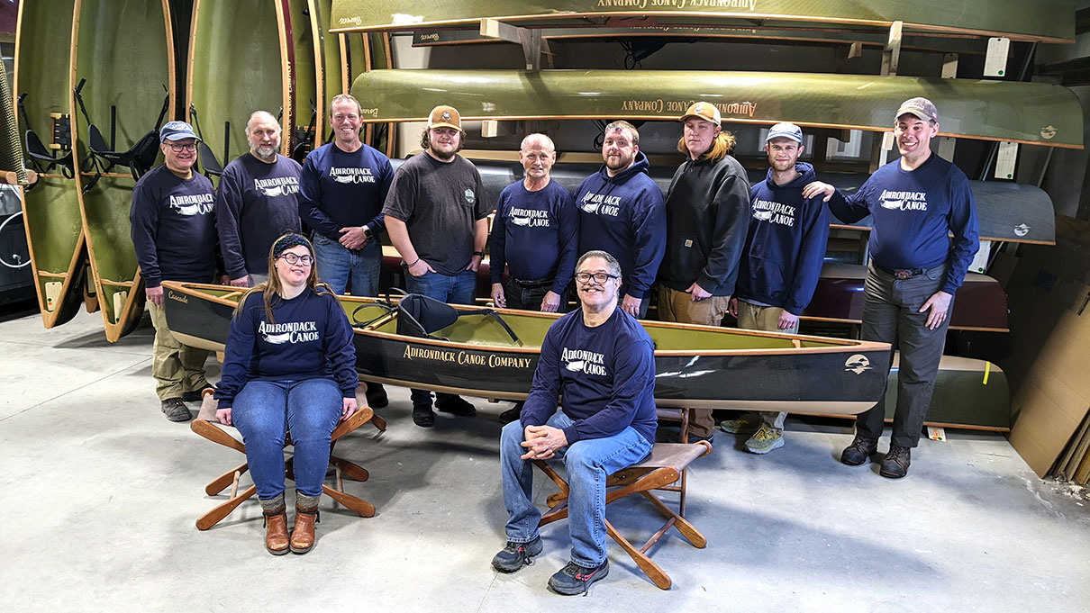 The crew at Adirondack Canoe Company poses in their warehouse in front of a rack of canoes