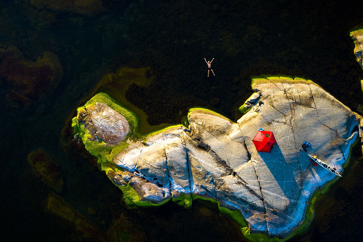 overhead photo of a small rocky island with campsite and kayak while person floats spread-eagle in water nearby