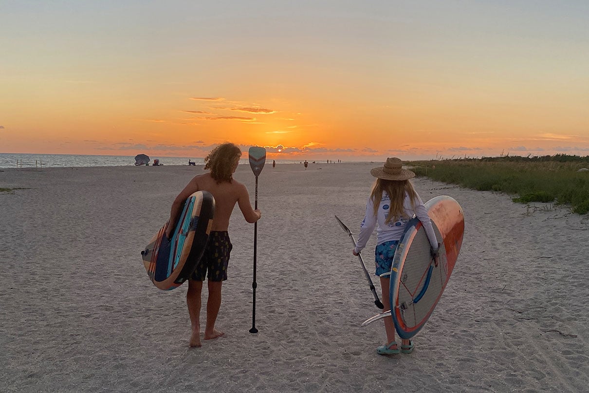 two paddleboarders walk along a scenic Florida beach at sunset while carrying their SUPs and paddles