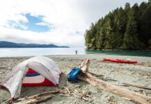 person stands on beach with arms outstretched at remote coastal campsite with kayak, tent and pack nearby