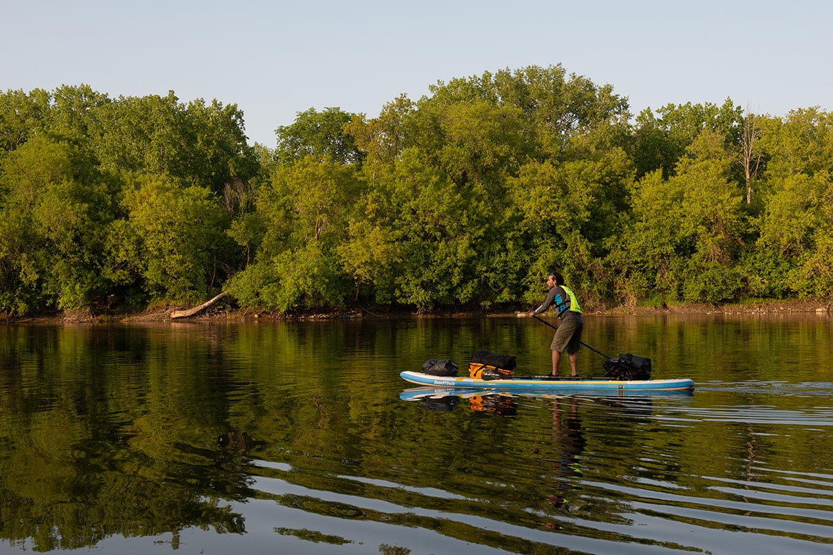 man standup paddleboarding past a leafy riverbank