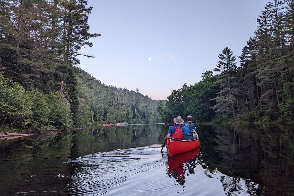 canoeists paddle along a river in daytime while the moon is up in the sky