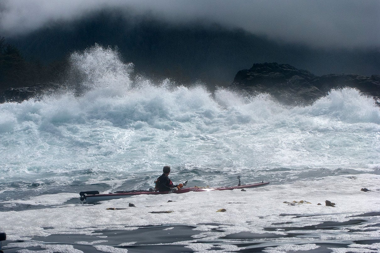 surf crashes over rocks with grey clouds in background and a sea kayaker in foreground