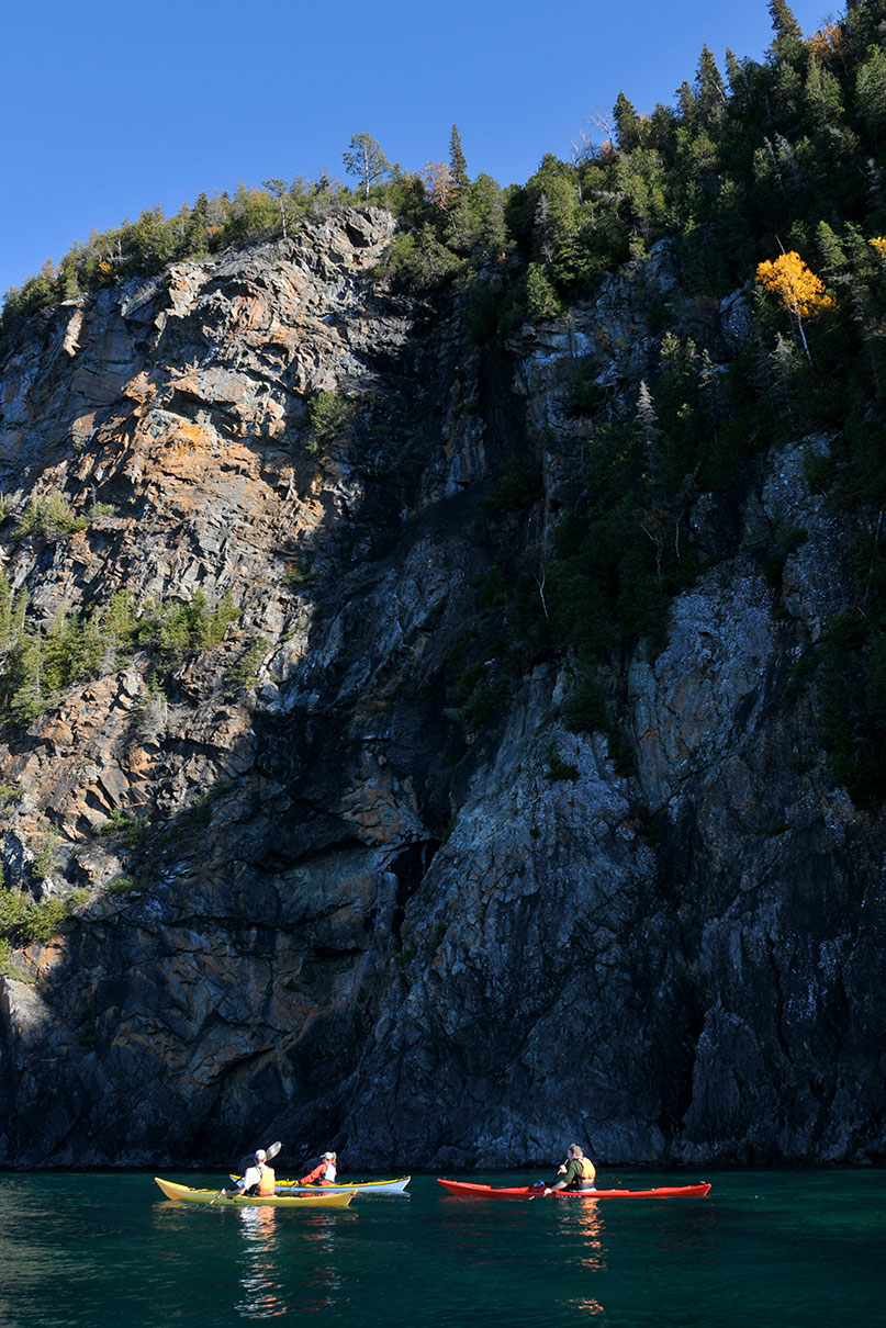 three sea kayakers paddle past the base of a dramatic rocky cliff