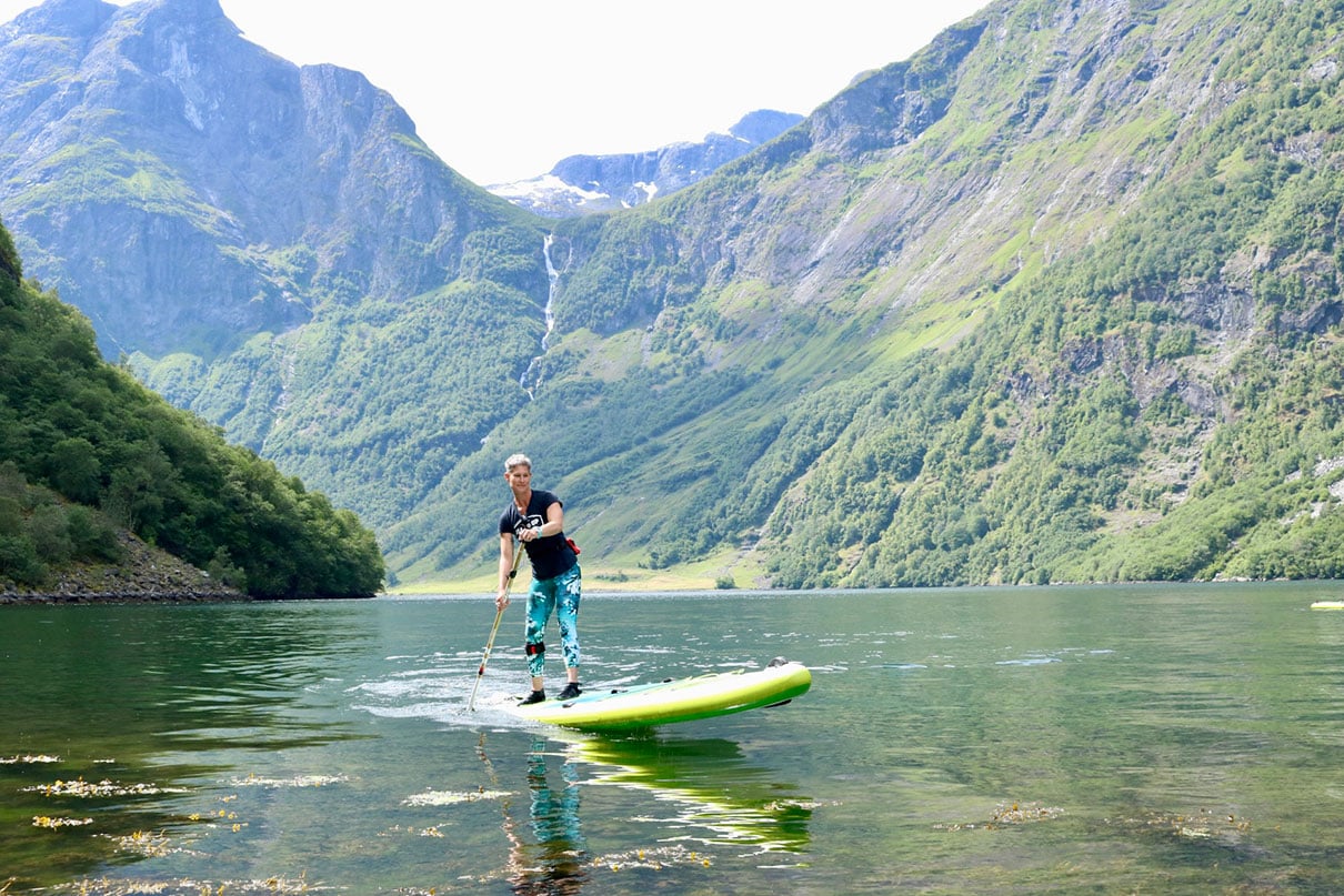 woman paddleboards in a Norway fjord with thin waterfall in background