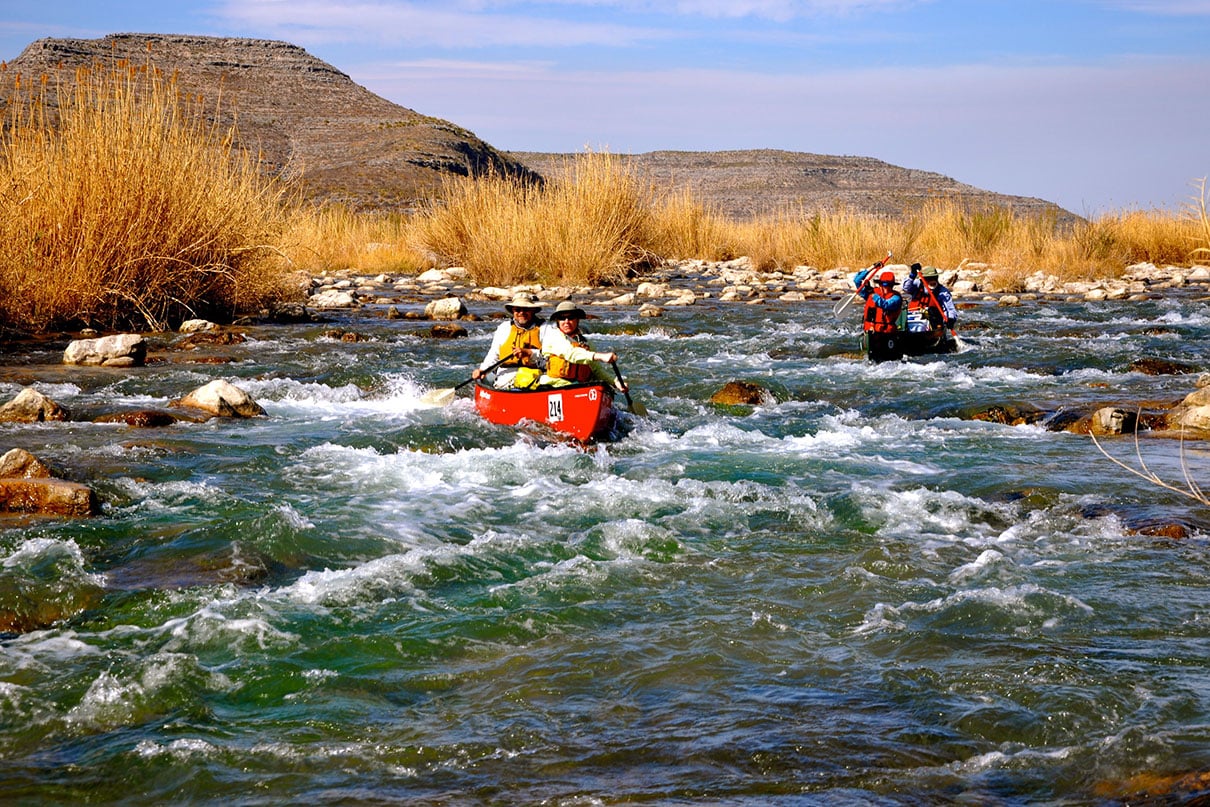 paddlers in two canoes make their way through light rapids with grass and hills in background